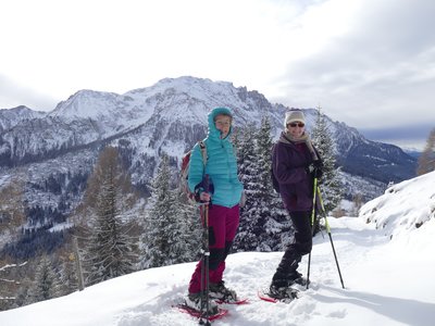 Two walkers wearing snowshoes and holding ski poles standing on edge of snow-covered hill facing camera with snow-covered mountain in background, St Zyprian, Italy