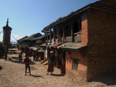 Dirt path lined with traditional stone and wooden houses with colourful fabrics hung outside in rural village. Children are standing in the foreground below clear blue sky, Nepal
