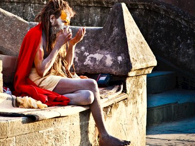 Holy sadhu man with painted face, sat with a folded leg at Pashupatinath Temple and blowing into hands, Kathmandu, Nepal