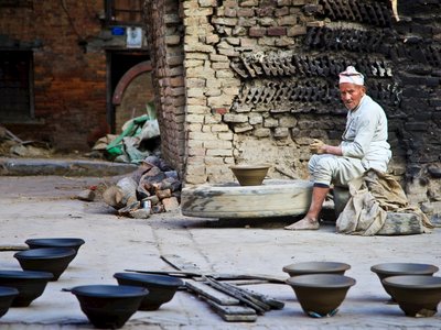 Man sat on chair at pottery workshop producing pottery with finished bowls drying in foreground, Nepal