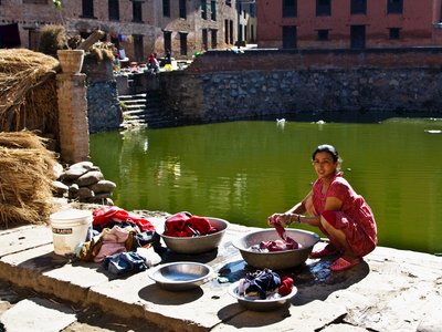 Woman in a red dress squats beside a green water, washing clothes in large bowls. Various articles of clothing are spread out on the ground, and the background features a brick building with visible steps leading to the water, Nepal