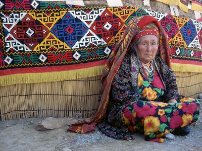 Traditional tapestry displayed over straw fence behind woman in colourfully patterned clothing, Kazakhstan