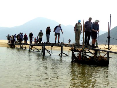 Ramble Worldwide walking holiday group crossing river by walking over thin wooden bridge close to water with mountains in background covered by mist, Hong Kong, Asia