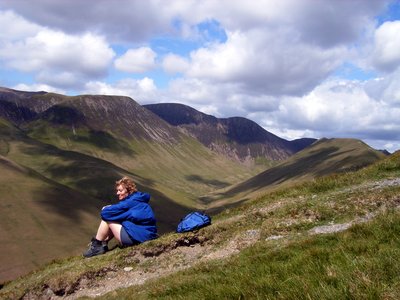 Lady on Ramble Worldwide walking holiday in outdoor clothing sitting atop of Lake District fell admiring view of peaks on sunny day with white clouds