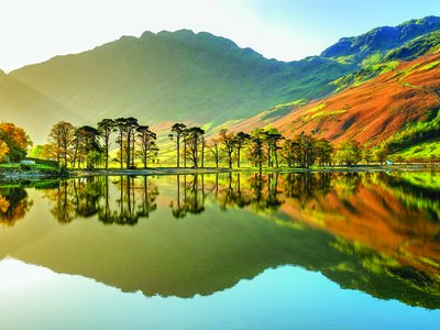 Buttermere Lake with reflections of colouring orange and green fells with trees and blue sky, Lake District