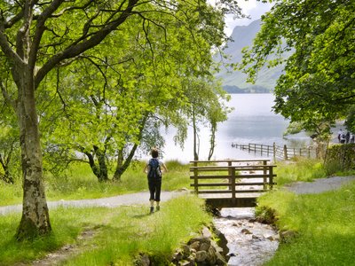 Lady with backpack standing next to stream beneath tree admiring Buttermere Lake on sunny day, Lake District