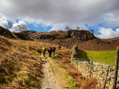 Ramble Worldwide walking holiday group walking in the distance along gravel path with rocky fells and farmland either side, Lake District