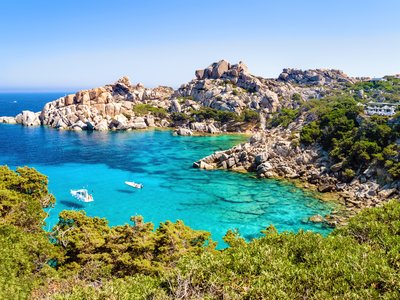 Two small boats floating in distance in turquoise water with coastal rocks and green trees formed around, italian island sardinia in mediterranean sea