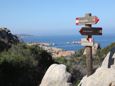Signposts showing direction of different walking trails in Sardinia with coast and sea view, with one stating "RISPETTA LA NATURA SEGUI IL SENTIERO"