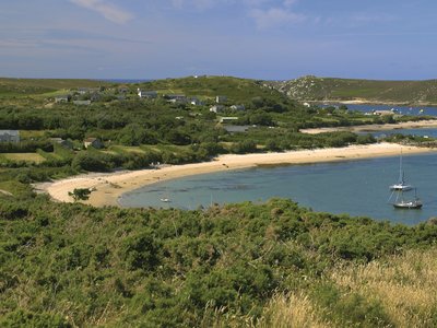 Curved beach edge of Bryher Island with green bushes growing over land and boats floating on water nearby, Isles of Scilly, England