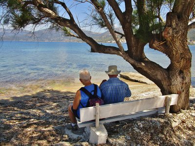 Man and woman sat on bench under shade of tree taking in view on Marmari beach, Kos, Greece