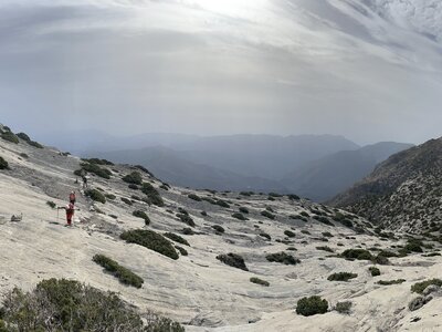 Group of hikers moving up smooth wavy chalk white mountain dotted in green shrubs with outlines of mountains hiding in background from clouds, Sierra de las Nieves, Spain