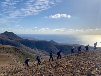 Diagonal line of people ascending hill from sideview in mountainous landscape with sun shining across sea in background, Lanzarote, Spain