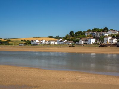 Peaceful village with white houses nestled along the water’s edge, set against rolling green and golden fields under a clear blue sky. Sandy banks line the calm river, and a few boats and seagulls are visible along the shore. Bideford, East-the-Water