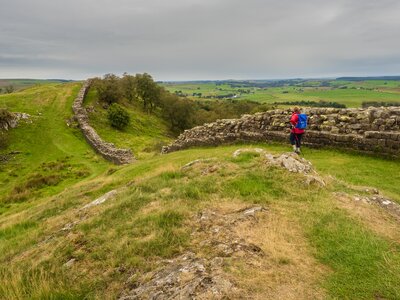 Female walker descending grassy slope alongside grey bricked Hadrian's wall in Northumberland, England