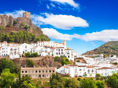 White and beige buildings nestled into hills of mountainous area with tall pine trees dotted throughout and a castle atop the highest point during sunny day, Zahara de la Sierra town, Sierra de Grazalema, Cadiz, Andalusia, Spain