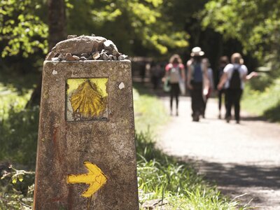 Way of St James walking marker with yellow seashell and right arrow beneath cement structure pointing towards walkers along path in distance, Camino de Santiago