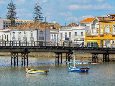 Tavira river with low bridge arching over towards bright white and yellow houses and small boats floating in foreground, Algarve, Portugal