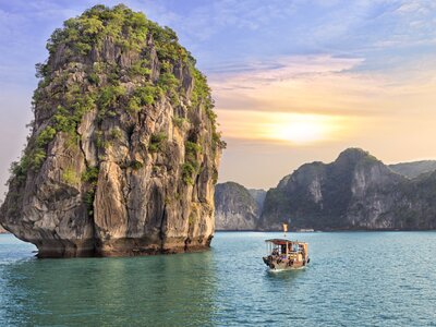 Large freestanding rock with green trees growing on edges and small boat nearly in body of water with sun glowing and mountainous peaks in background, Ha Long Bay, Vietnam