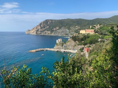 Monterosso al Mare town viewed from distance atop hill with trees in foreground and coastal mountainous landscape close to sea on sunny day, Italy