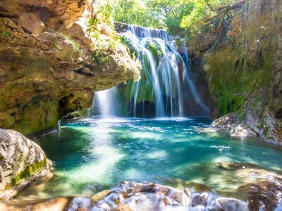 Long-exposure shot of Waterfall of Akchour with sun spots appearing through tree canopy on surface of turqoise water and tall rocks surrounding, Talassemtane National Park, Morocco