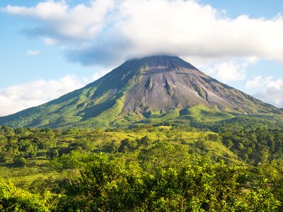 Arenal volcano seen in distance beyond luscious green trees with cloud floating directly above volcano, Costa Rica, Central America