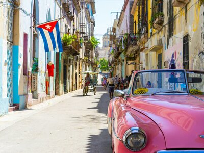 Pink 1950's Cadillac Chevrolet parked in street with cuba flag hanging on building with pedestrians walking nearby, Cuba