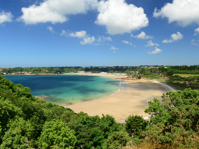 Sunny picturesque view of beach with clear blue waters and green trees and grass surrounding, Jersey, Channel Islands, UK