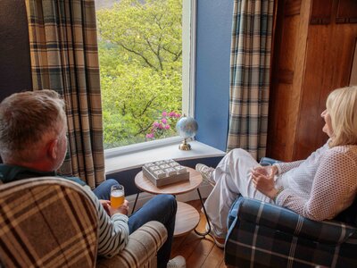 Two people sat in tartan pattern chairs by large window enjoying view of nature with beverages in hand