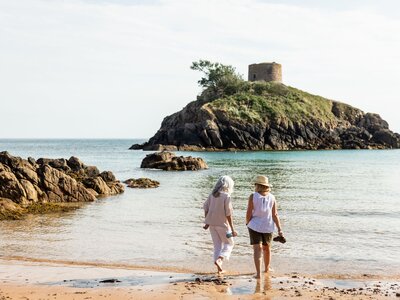 Couple stepping into water from sandy beach with small fort in distance, Portelet Jersey