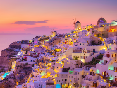 Oia village during sunset with pink and purple pastel colours across the sky and artificial yellow and blue lights rising from white buildings, Santorini, Greece