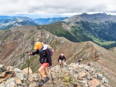 People ascending on hike along rocky mountain ridge to the Pic de Sanfonts with large mountainous terrain in background on cloudy day, Andorra
