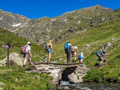 Walking group on Ramble Worldwide walking holiday enjoying sunny day in Andorra coming from Estanu Blau and crossing over small wooden footbridge with water flowing beneath and signpost nearby with green rocky mountain landscape in background