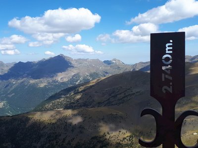 View from Pico Casamanya south peak with signage in foreground showing 2.740m. hollow metallic engraving with mountain range in background showing fluffy white clouds and their shadows casting onto mountains