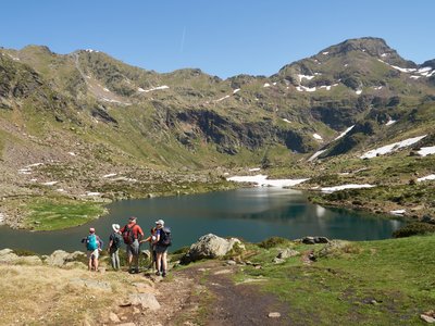 Ramble Worldwide walking group congregating towards bottom of footpath admiring Tristaina Lakes with mountainous landscape and snow melting exposing green grass on sunny day, Andorra
