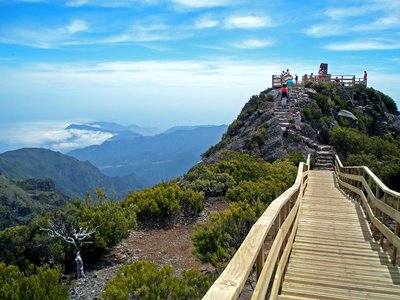 Wooden decking and stone pathways leading towards people standing at viewpoint at Pico da Ruivo with vast mountain landscape spanning into far distance, Madeira, Portugal