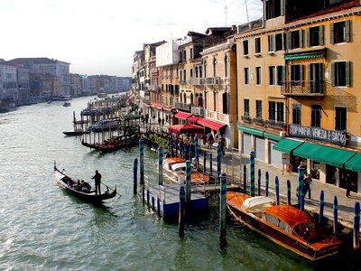 High view of boats docked by path and row of shops on canal edge, Venice, Italy