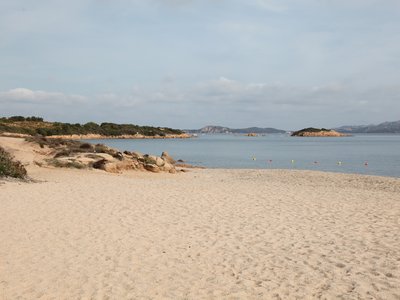 Sandy beach with scattered rocks and low vegetation, leading to calm blue sea with small islands and distant hills under a partly cloudy sky, Sardinia, Italian island in the Mediterranean Sea